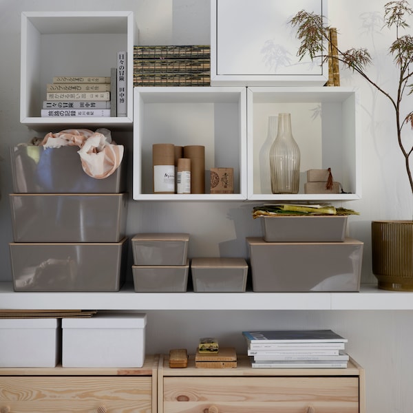 Several dark grey and beige KUGGIS boxes in different sizes are placed on a wall shelf with 4 white EKET cabinets above.